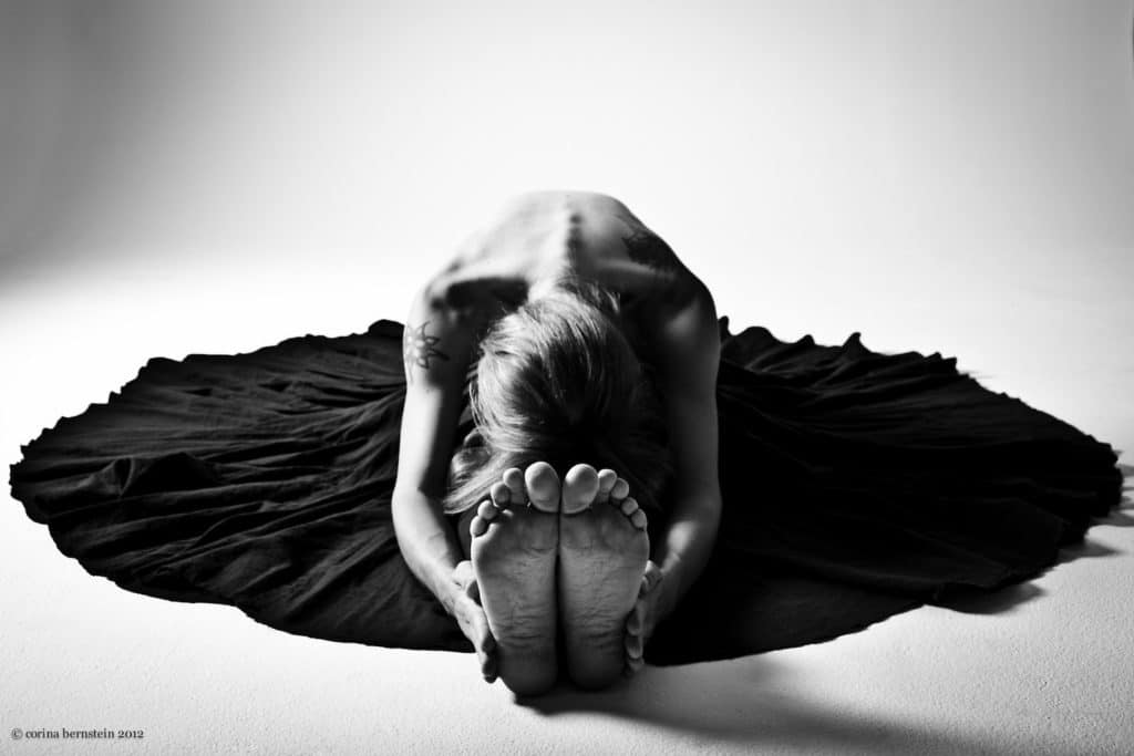 black and white image of a women in a black dressed, sitting on the floor, reach to her toes toward the camera 