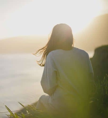 A photograph of a woman standing on a mountain, gazing towards a picturesque beach below, as the sunrise creates a beautiful halo around her head.