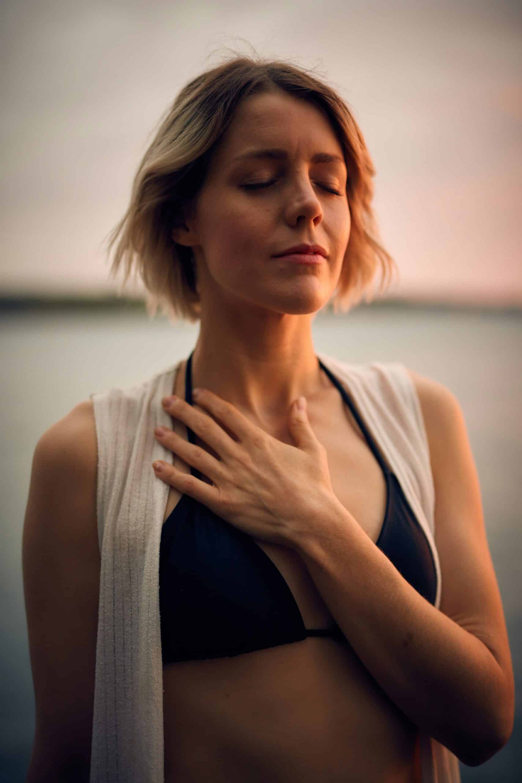 Woman stands, eyes closed, facing the setting sun and placing her hand on her chest as she breaths deeply
