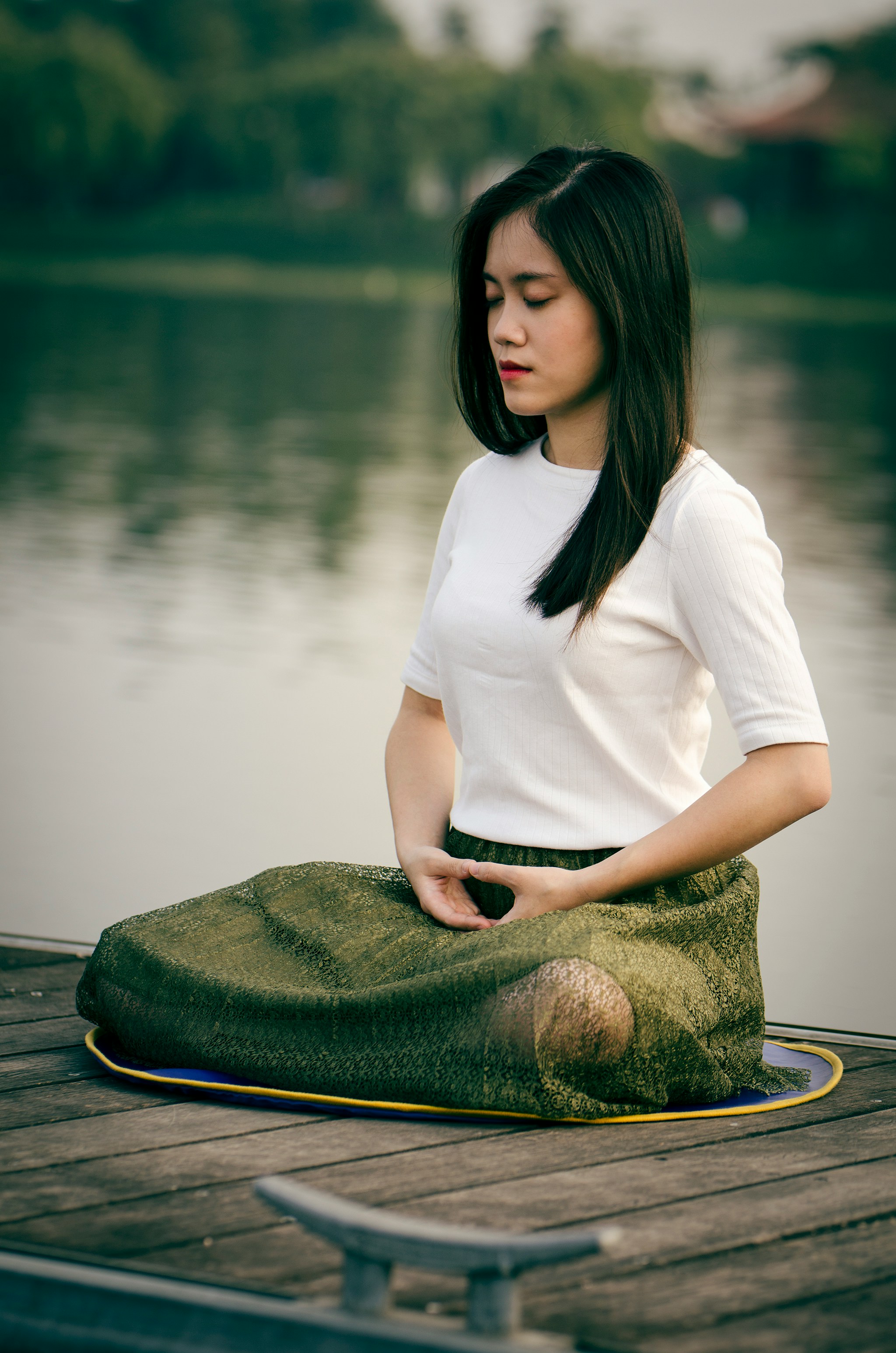 A woman sits cross legged on a dock by a lake with her eyes closed as she practices somatic breathing