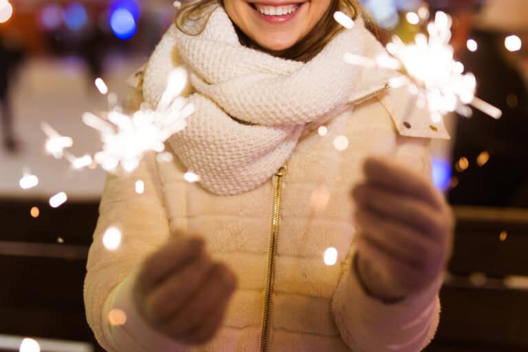 A person wearing a white coat, scarf, and gloves is holding two sparklers in their hands, smiling in a festive, outdoor winter setting with blurred lights in the background.